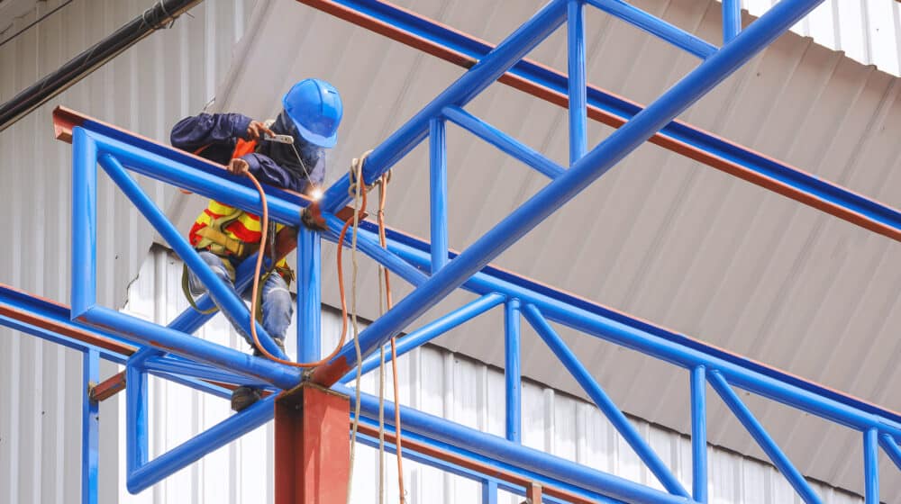 Construction worker with safety equipment is welding metal roof structure of warehouse building in construction site